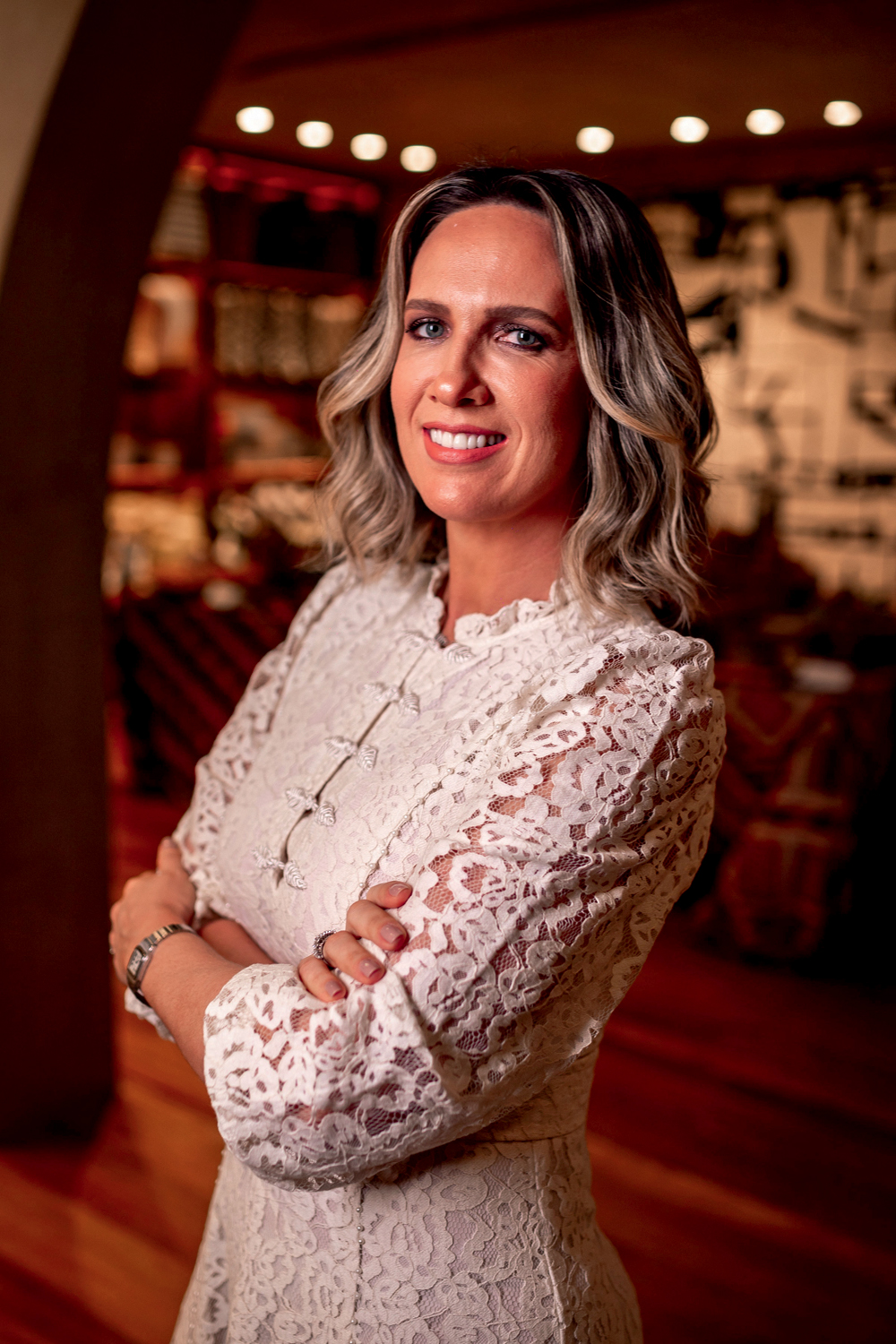 Mulher loira posa com vestido branco de braços cruzados e sorrindo