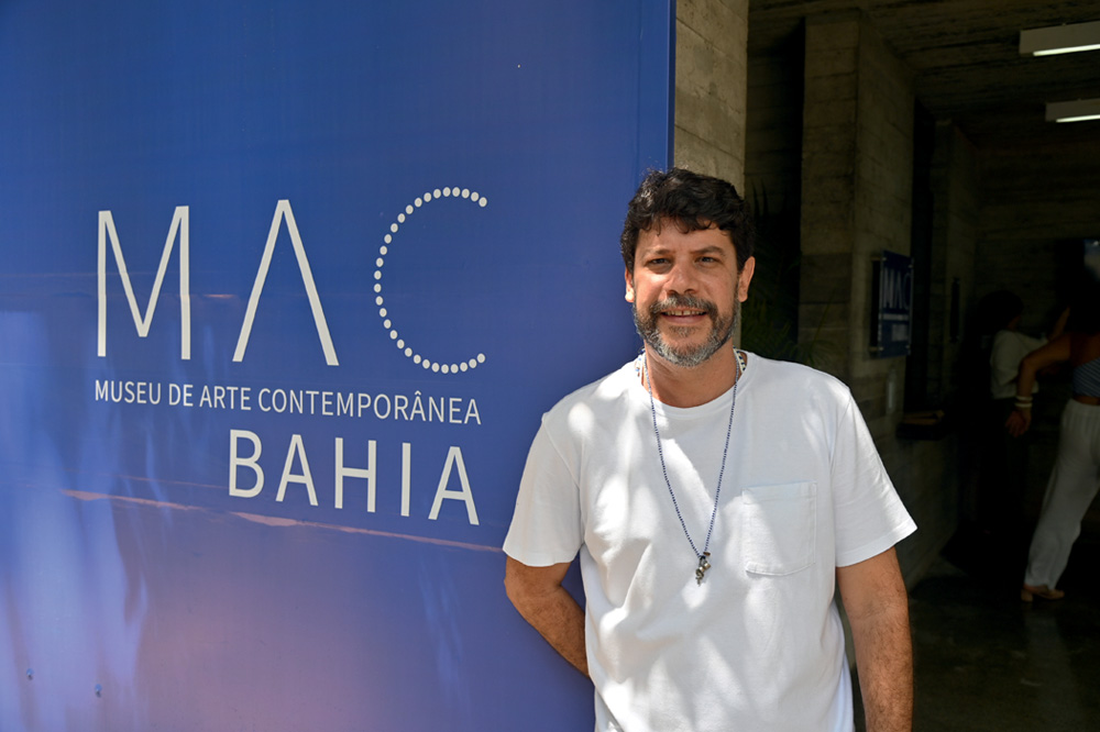 Homem branco de camiseta branco ao lado de painel azul indicando MAC Bahia