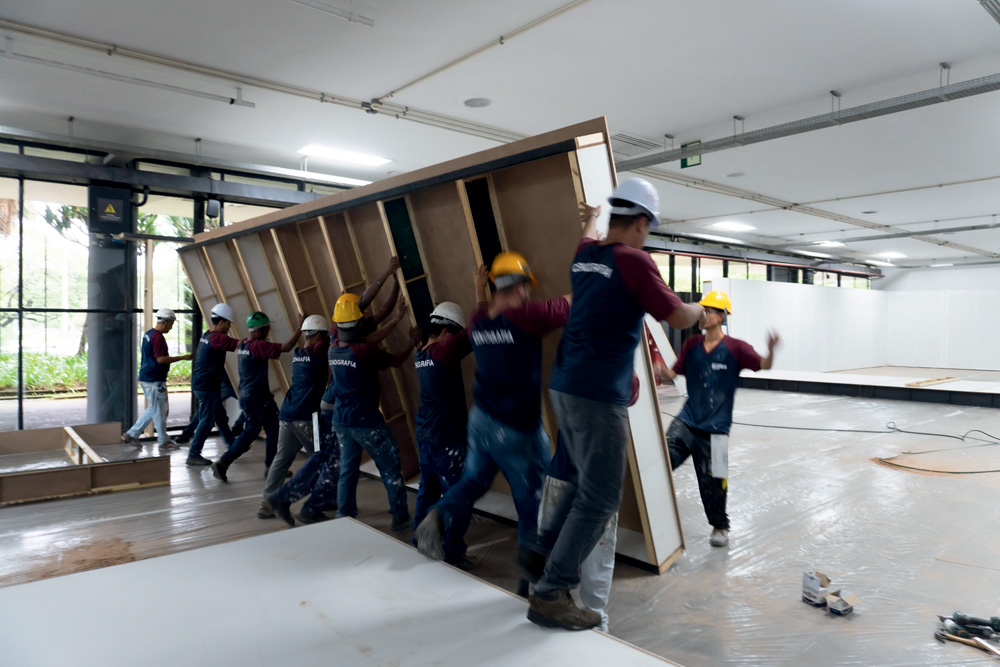 Homens erguem peça grande de madeira no meio de um pavilhão