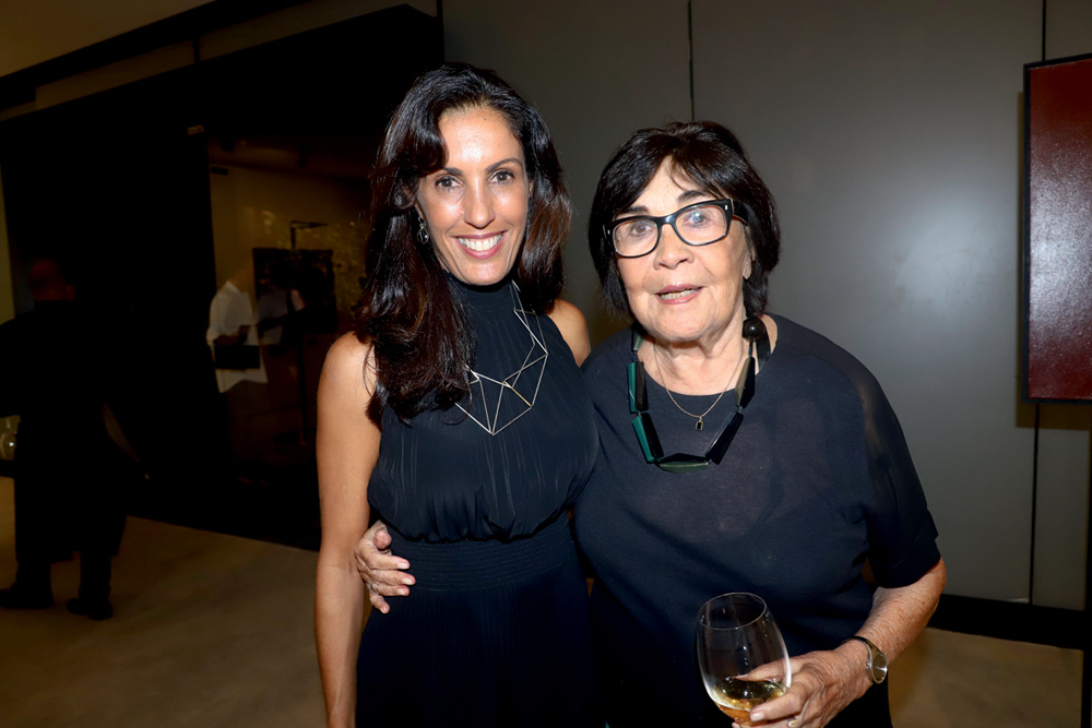 Duas mulheres sorriem, ambas vestindo peças pretas, a da esquerda com cabelos longos pretos, a da direita curtos