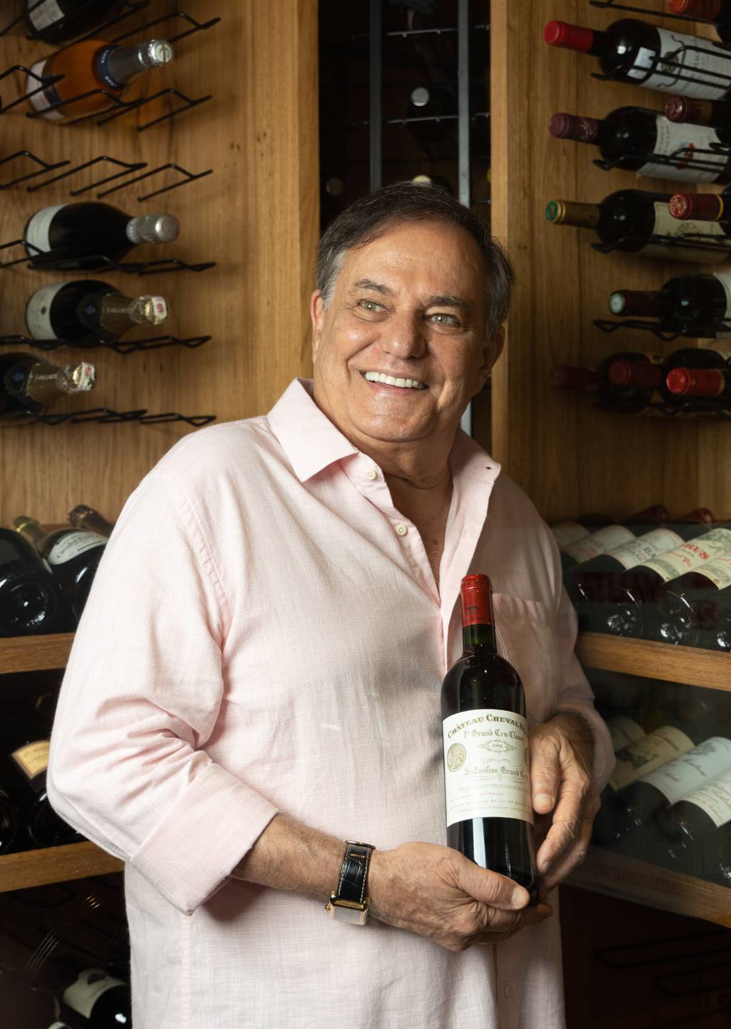 Senhor branco de cabelos grisalhos sorri olhando à esquerda e segurando garrafa de vinho. Veste camisa bege e posa em frente a prateleiras de madeira contendo fileiras de garrafas