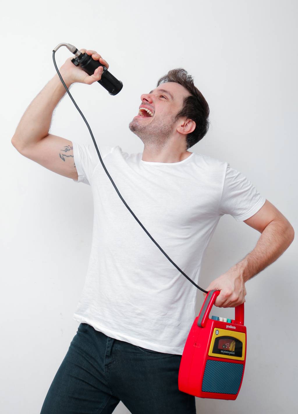 Homem branco posa fingindo cantar com microfone de brinquedo, uma caixa de som vermelha à mão e a outra erguida com o microfone no alto