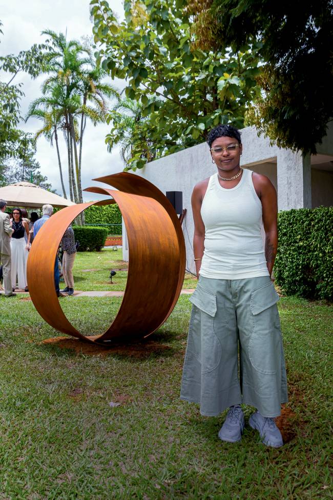 Imagem mostra Siwaju em pé em gramado, com grande obra de arte em formato circular ao fundo