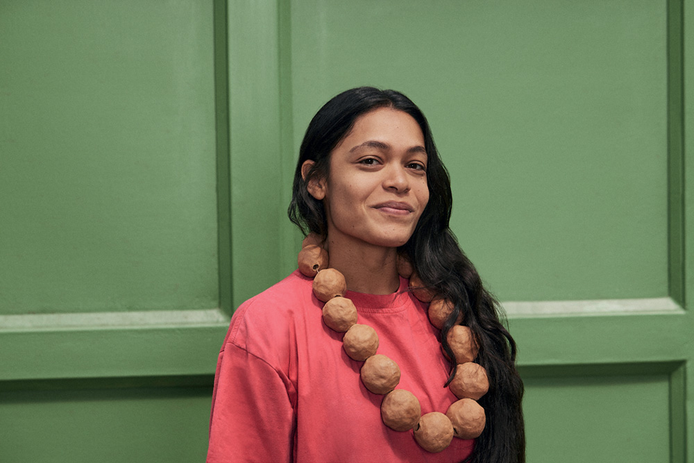 Mulher indígena de cabelos pretos longos sorri olhando para a foto, usa colar grande de esferas marrons e usa peça rosa