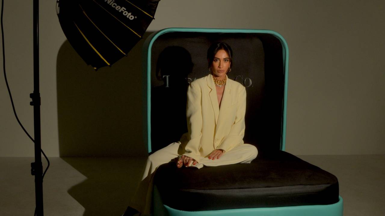 Mulher posa sentada em sofá em formato de estojo de joia vestindo roupas brancas dentro de um estúdio