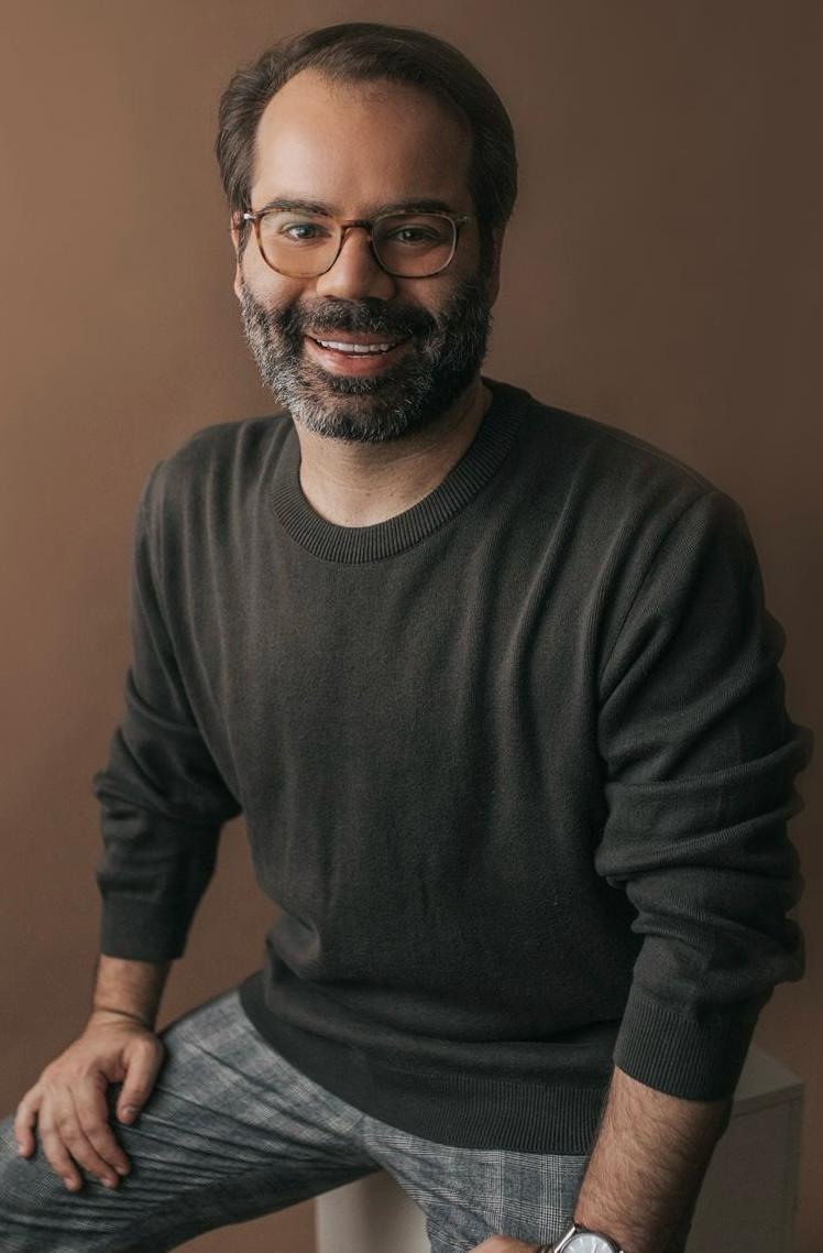 Homem de óculos redondo e barba com pelos brancos e pretos posa sorrindo em fundo marrom e vestindo leve suéter preto