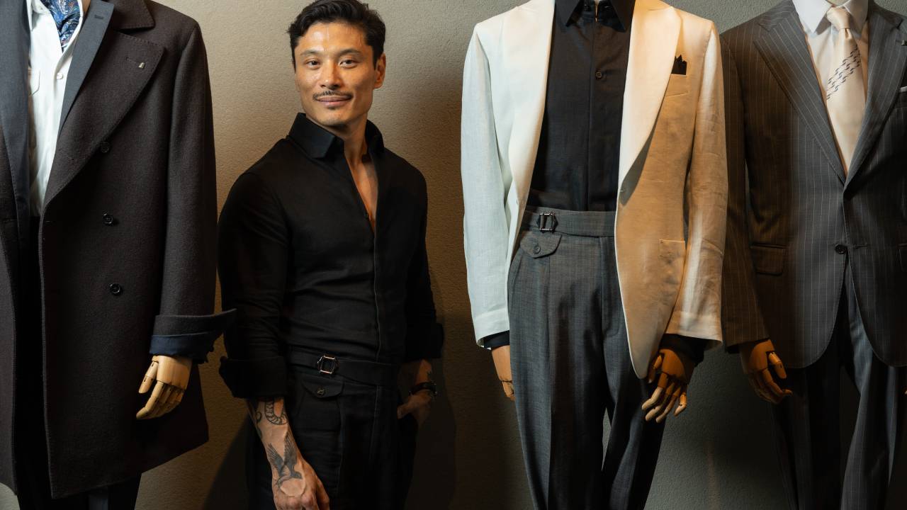 Homem asiático posa de pé entre manequins de ternos. Veste camisa e calça pretas