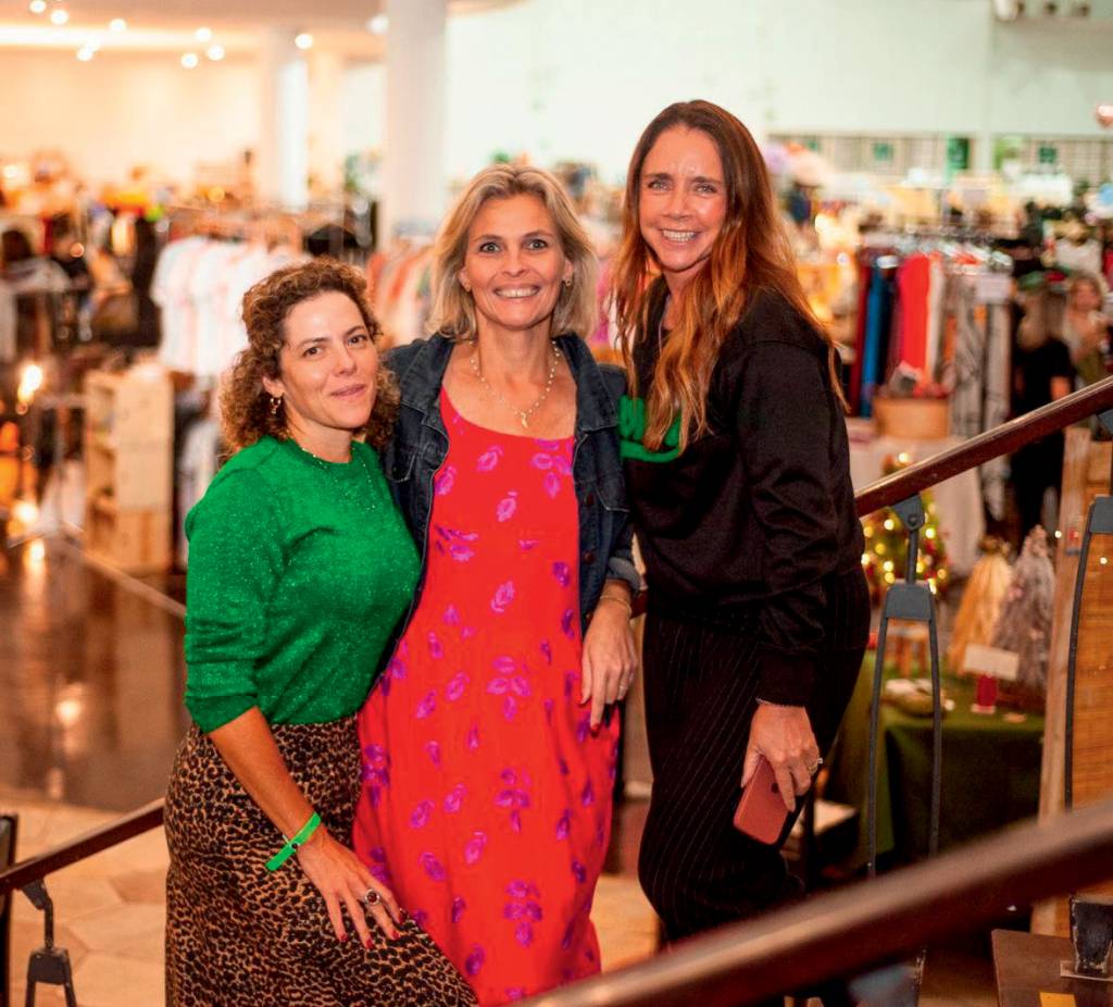 Três mulheres posam lado a lado em loja. A partir da esquerda, vestem peça verde, rosa e preta. As três sorriem.