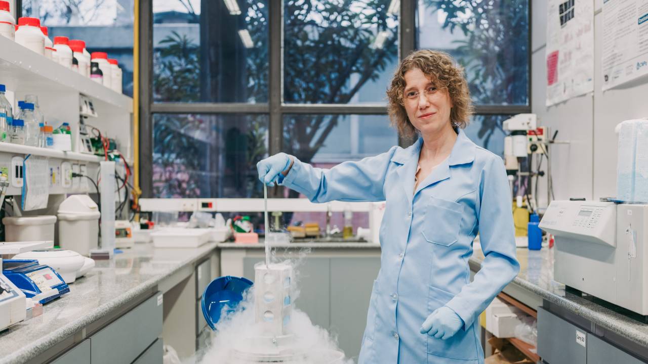 Mulher cientista de cabelos loiros encaracolados posa com avental azul segurando utensílio que sai de um copo fumegante no laboratório