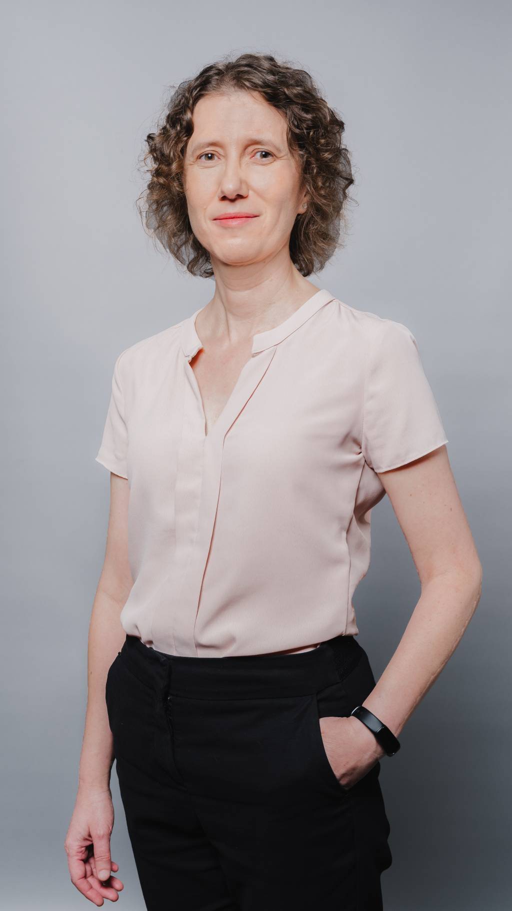 Mulher cientista de cabelos loiros encaracolados posa com blusa bege e calça preta com uma mão no bolso
