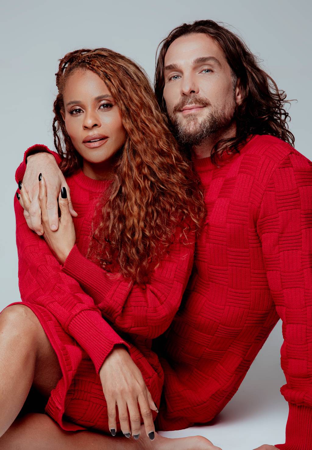 Mulher negra e homem branco, ambos de cabelos longos, posam sentados lado a lado, ele com o braço no ombro dela. Ambos vestem suéter vermelho