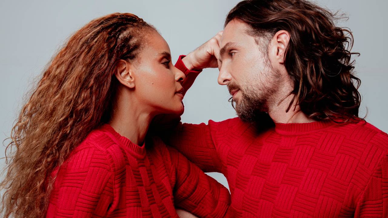 Mulher negra e homem branco, ambos de cabelos longos, se encaram de perto. Ambos vestem suéter vermelho
