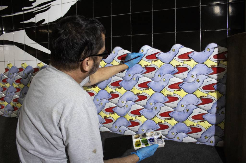 Foto exibe homem de costas restaurando mural de azulejos em corredor do metrô