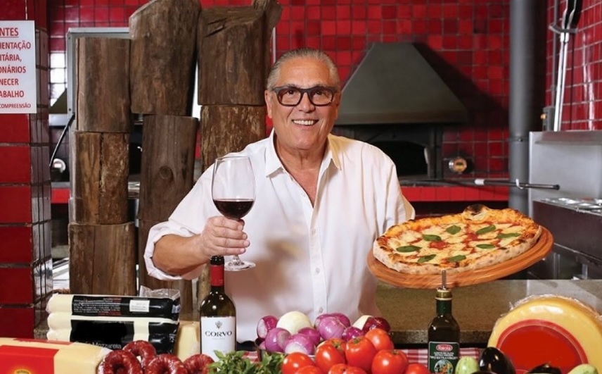 Um homem grisalho e sorridente com taça de vinho em uma mão e uma pizza na outra