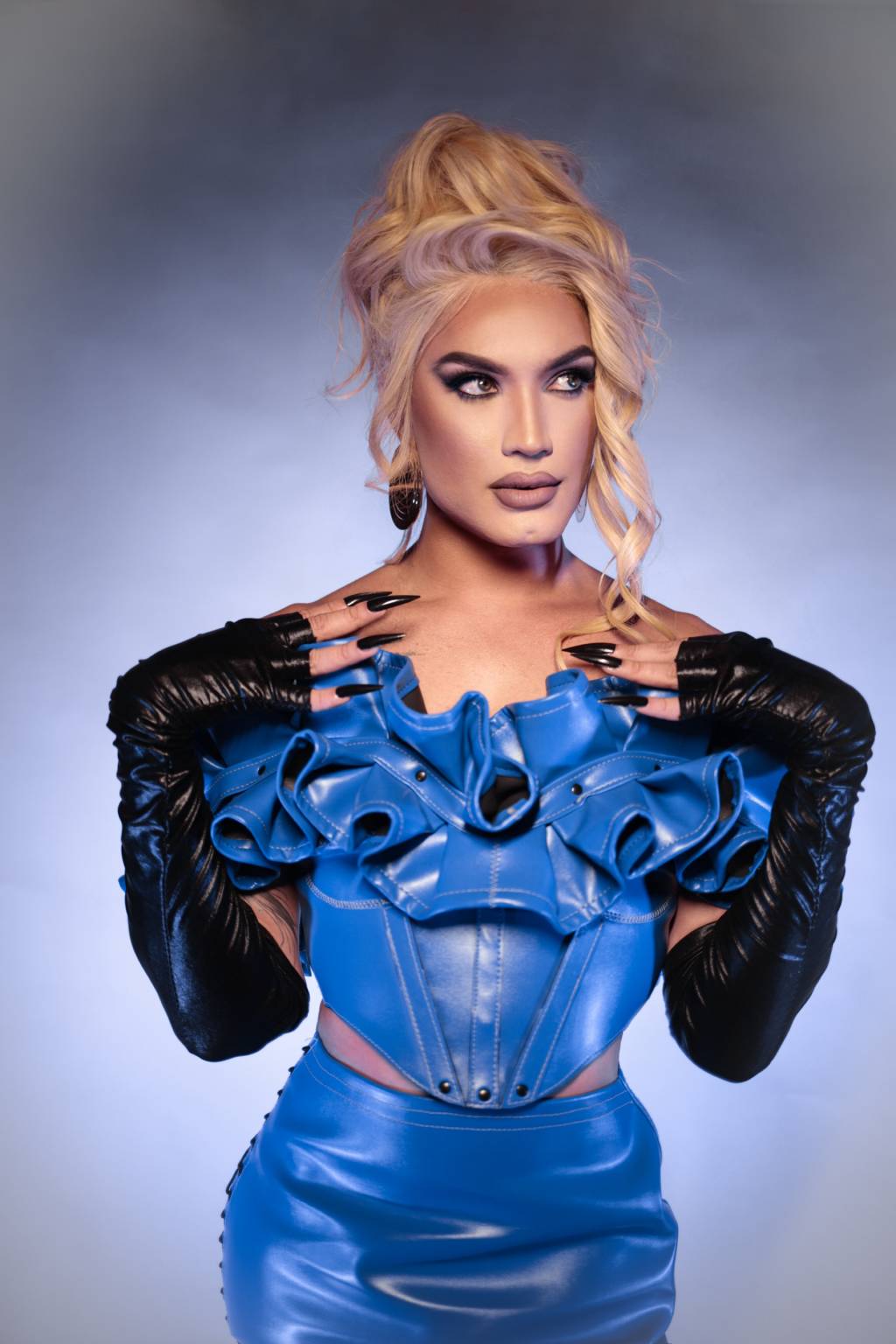 Aretuza Lovi-Crédito Maria Paula Freire-Divulgação (3) Drag queen loira veste traje azul com aparência de látex e babados na área do peito, além de luvas longas pretas.