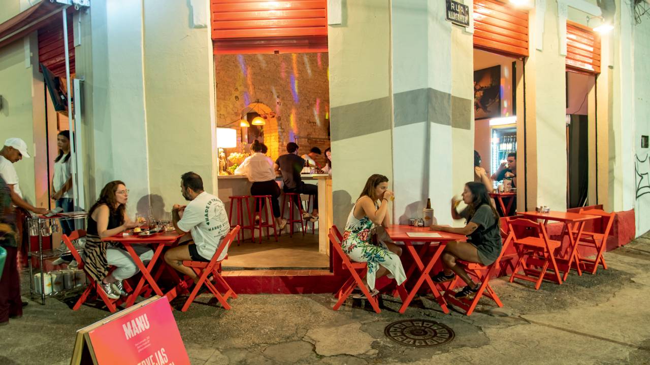 Fachada de bar com paredes creme e mesas vermelhas na calçada ocupadas por pessoas.