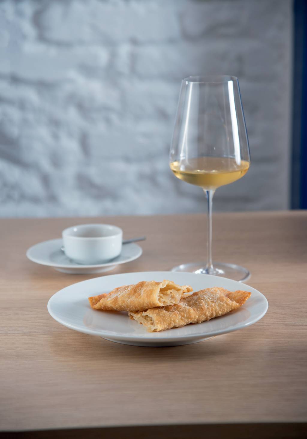 Prato de louça branca com dois pastéis retangulares sobre mesa de mandeira em frente a taça com vinho branco