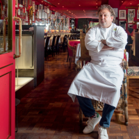 Chef branco de dólmã e braços cruzados sentado em poltrona em frente a salão de restaurante com paredes vermelhas cheias de quadros.