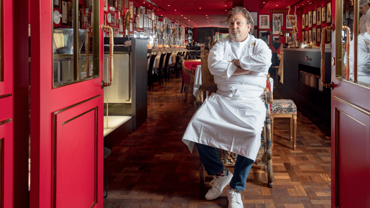 Chef branco de dólmã e braços cruzados sentado em poltrona em frente a salão de restaurante com paredes vermelhas cheias de quadros.