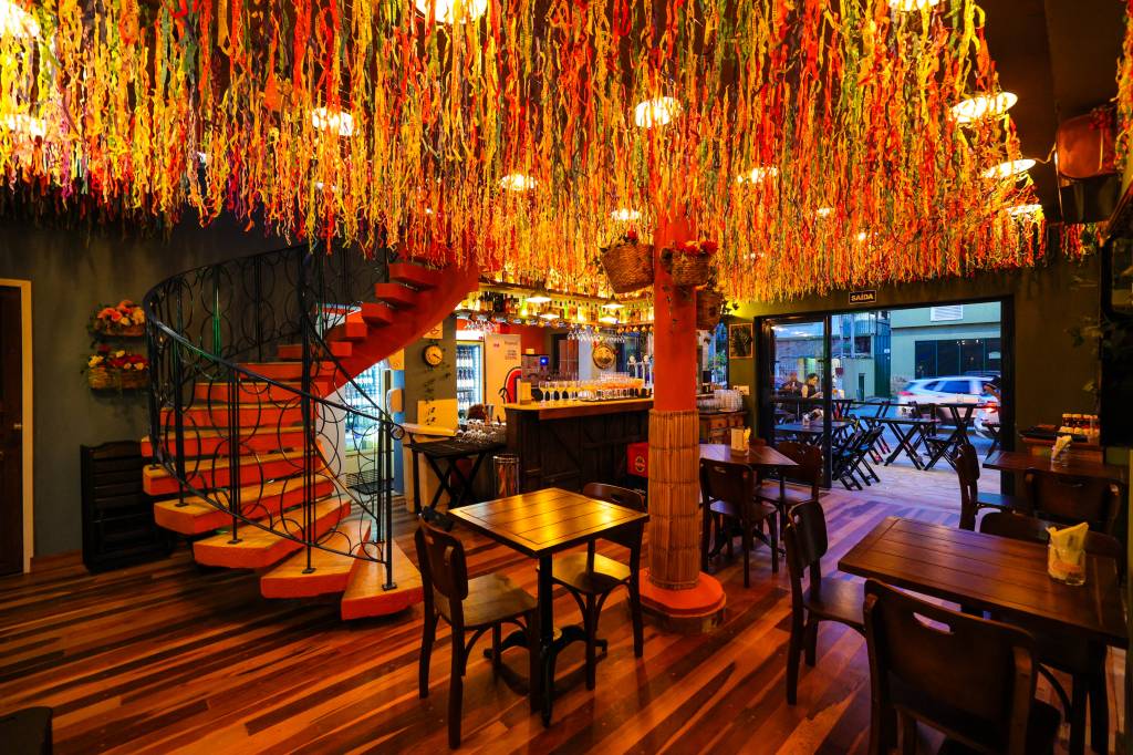 Salão do bar Tatuzinho com fitas coloridas presas no teto, chão e mesa de madeira e escada à esquerda.