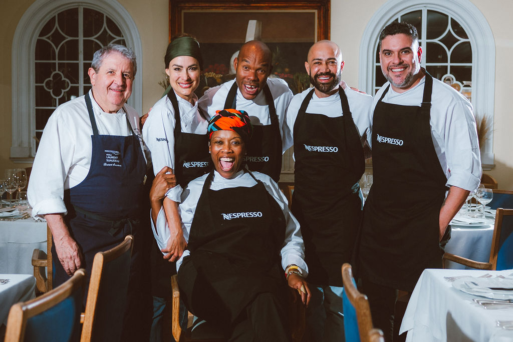 Seis chefs de dólmã branco e avental preto da Nespresso sorrindo para a foto. Cinco em pé atrás e uma chef sentada à frente.