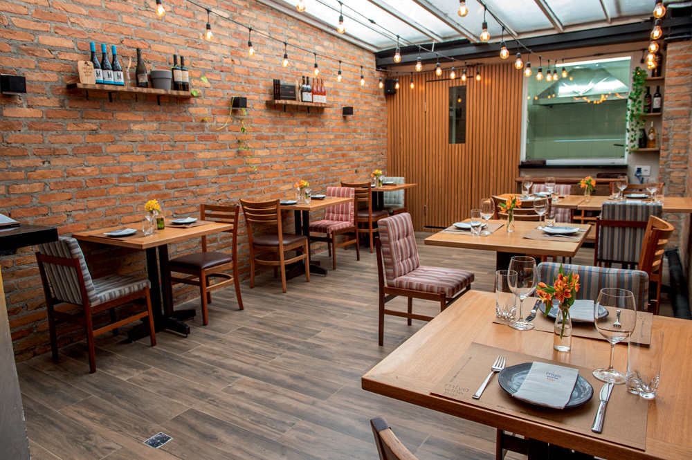 Salão de bar com mesas e chão de madeira, parede de tijolo aparente à esquerda e luzinhas no teto transparente.