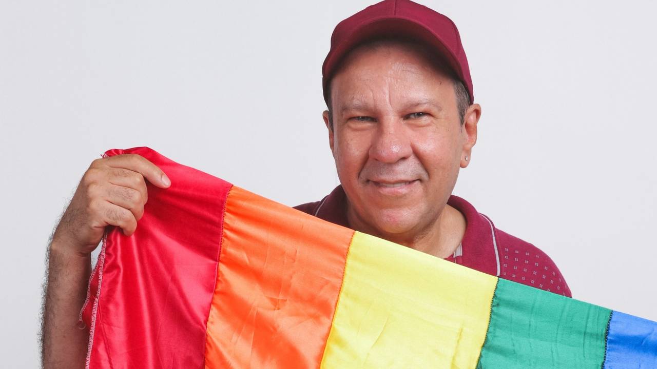 Homem branco de boné vermelho segura bandeira com as cores do arco-íris