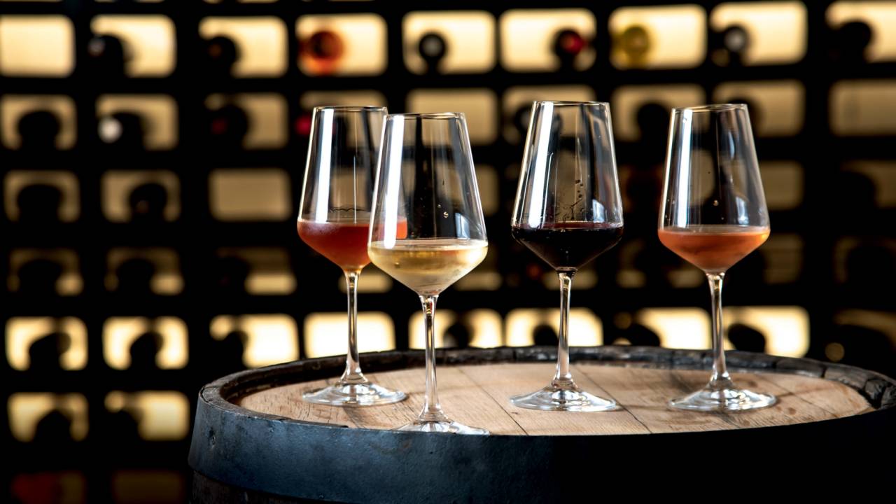 Quatro taças de vinho com vinhos de cores diferentes dentro sobre barril de madeira com adega ao fundo.