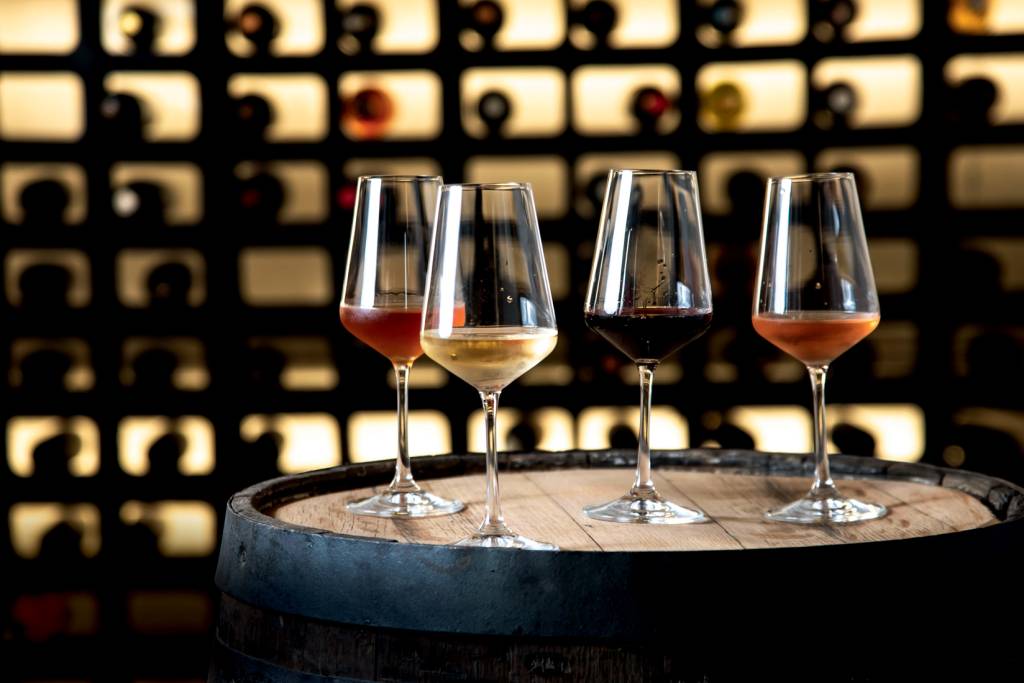 Quatro taças de vinho com vinhos de cores diferentes dentro sobre barril de madeira com adega ao fundo.