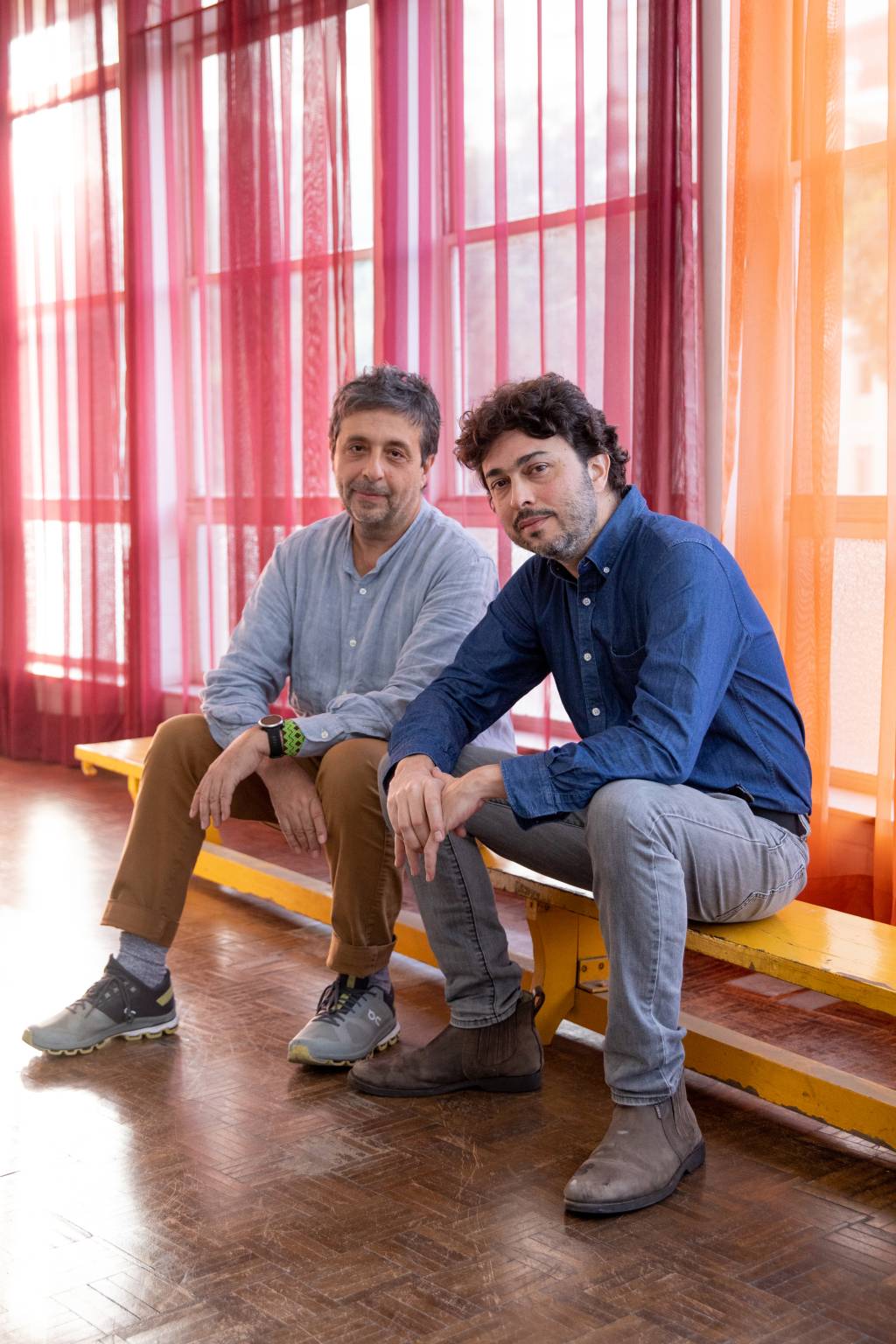 Dois homens brancos posam sentados em frente a cortina vermelha