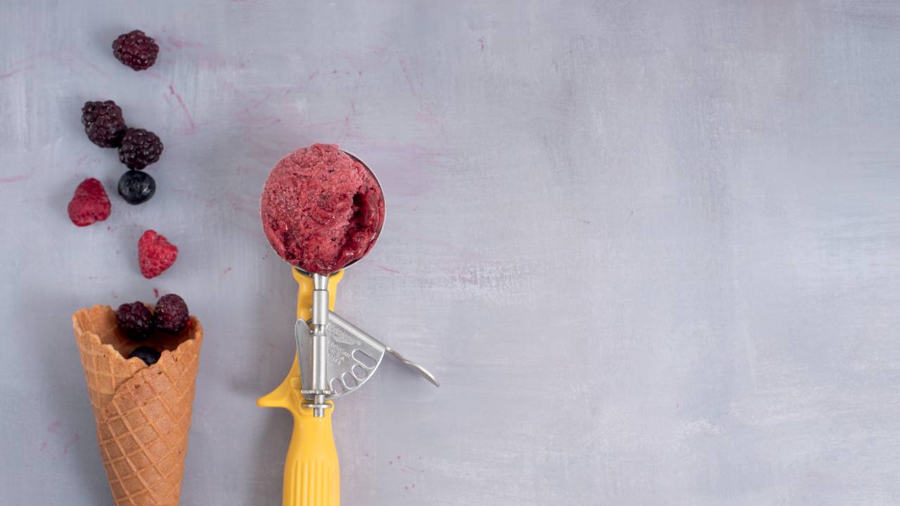 Foto aérea de colher de sorvete com bola de sorvete vinho ao lado de casquinha com frutas vermelhas dentro.