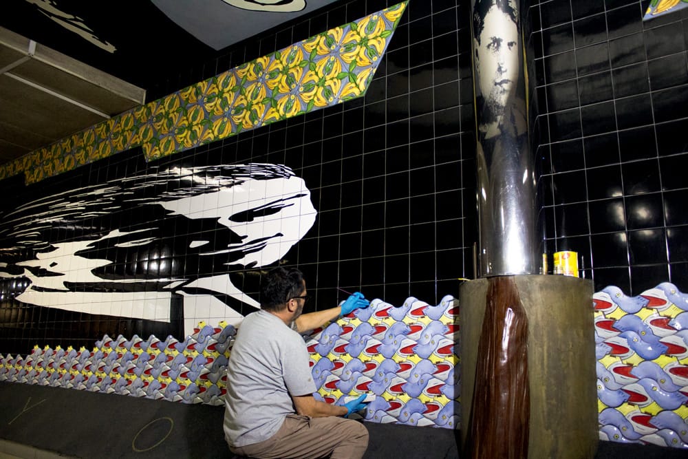 Foto exibe homem de costas restaurando mural de azulejos em corredor do metrô