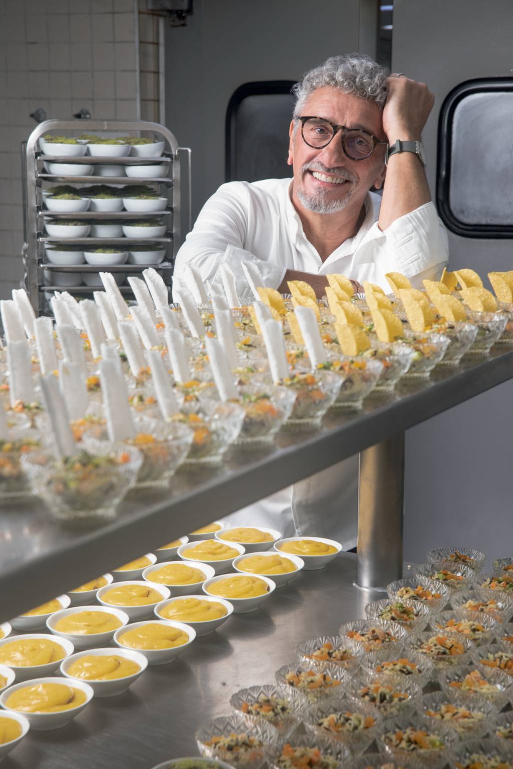 Chef sorrindo para foto com braços apoiados em prateleira metálica linha de produção de prato em cozinha industrial