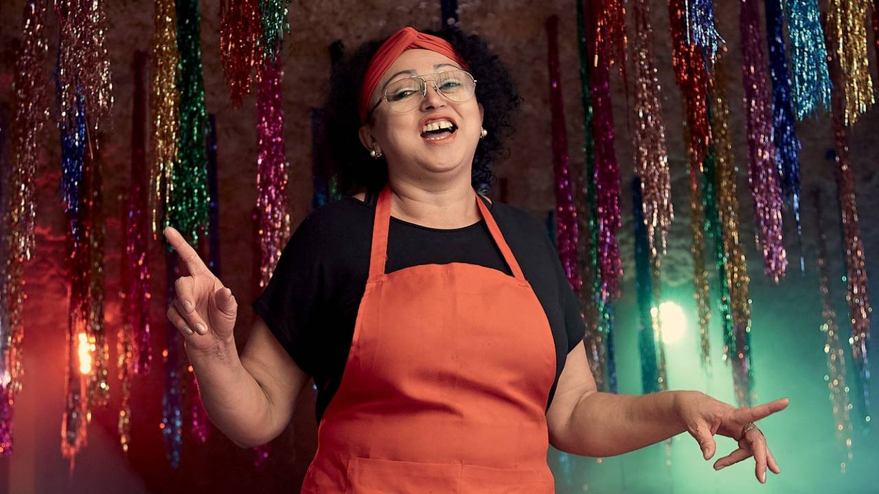 Moça de avental vermelho e pano vermelho na cabeça aparece dançando, em cima folhinhas coloridas e bola disco brilham