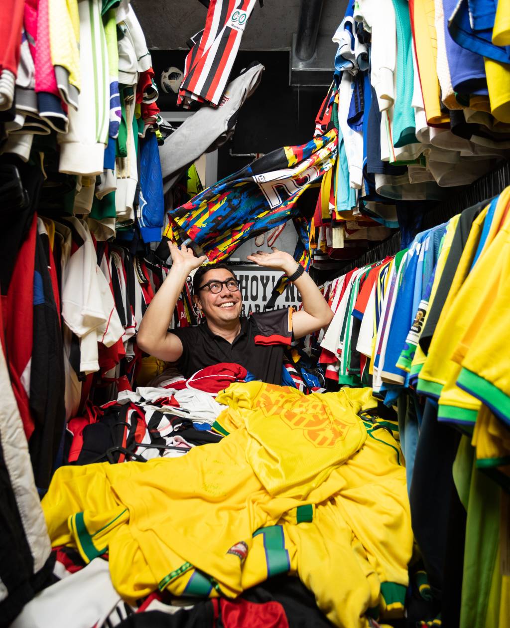 CASSIOCOLECCAMISAS22 Homem de óculos posa coberto por montanha de camisas de futebol, algumas em movimento caindo do teto