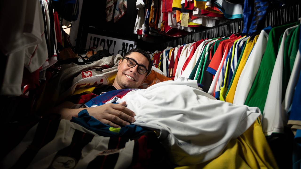 Homem de óculos posa coberto por montanha de camisas de futebol