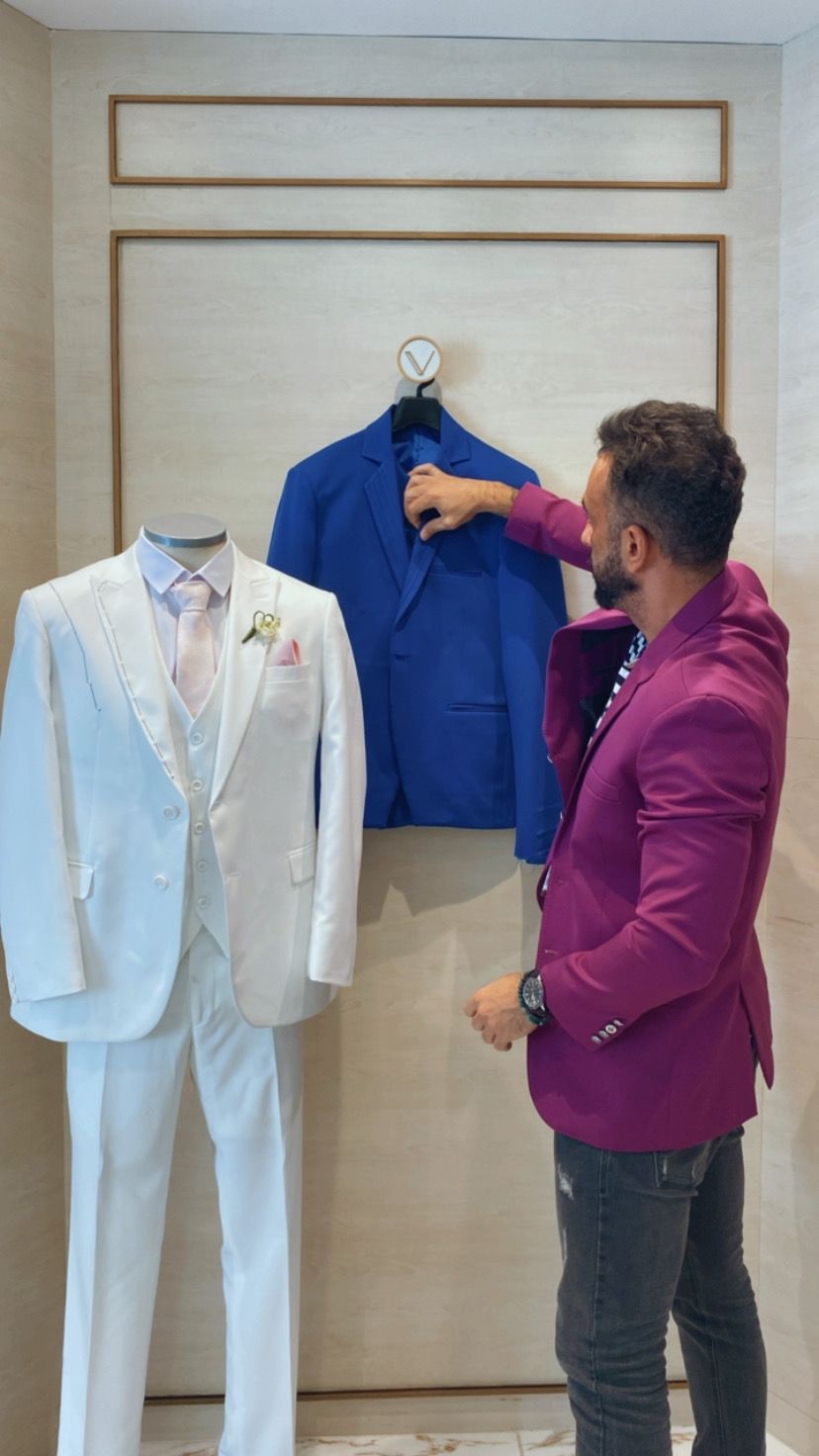 Homem de terno roxo mexe em paletó azul pendurado na parede. À esquerda tem um smoking completo branco em exposição
