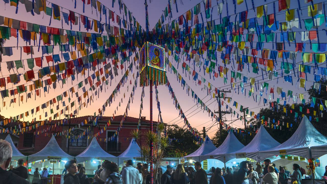 Festa Junina no final de tarde com bandeirinhas coloridas presas ao centro e descendo para diferentes lados. Ao fundo barraquinhas branca.