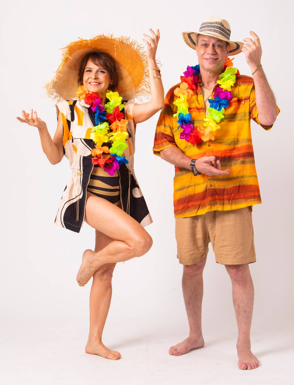 Casal hétero branco posa com roupas de férias de praia, com colar colorido e chapéus