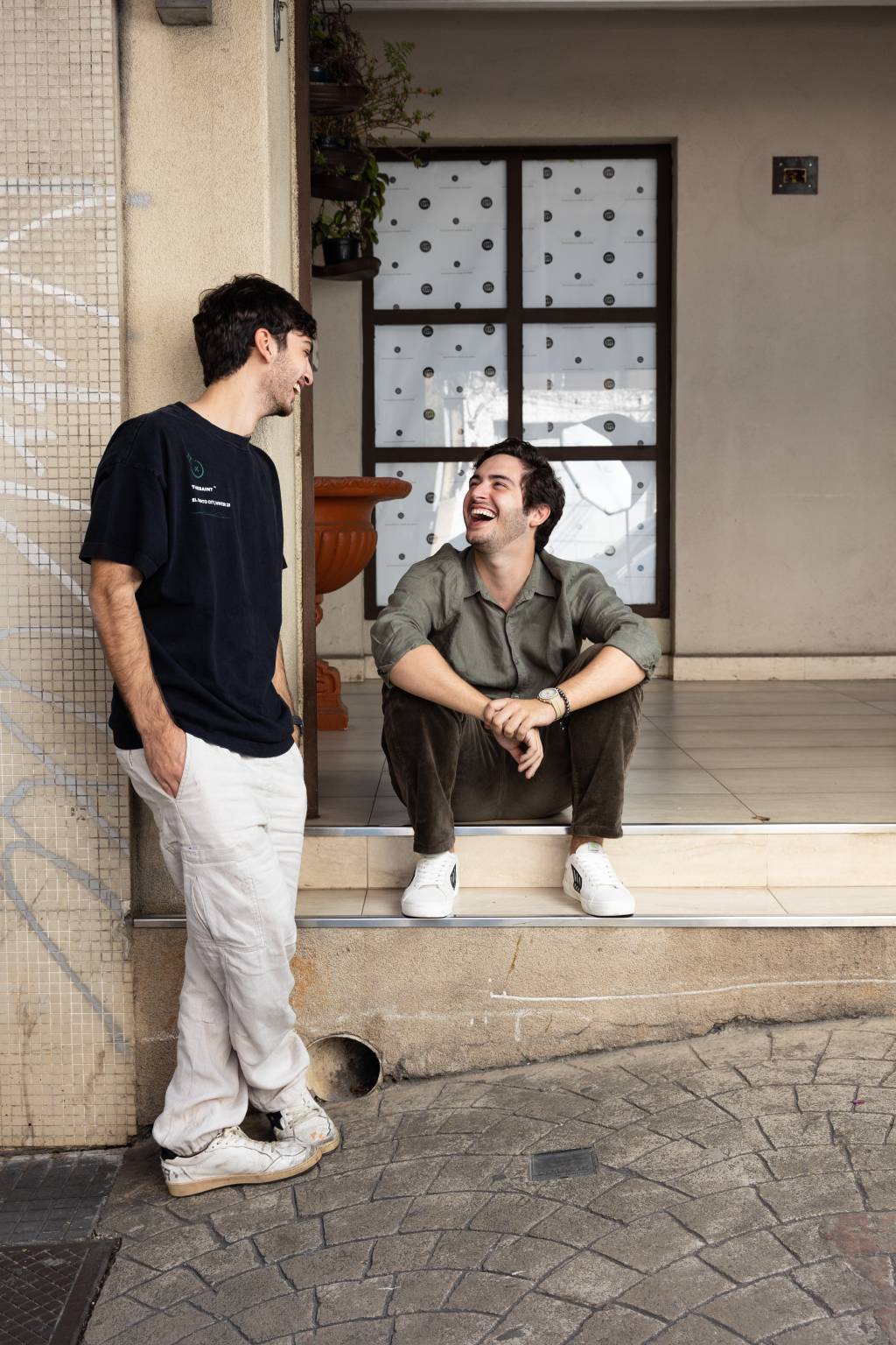 Dois jovens brancos posam encostados na fachada de galeria, um sentado e outro de pé. Eles riem e estão se olhando