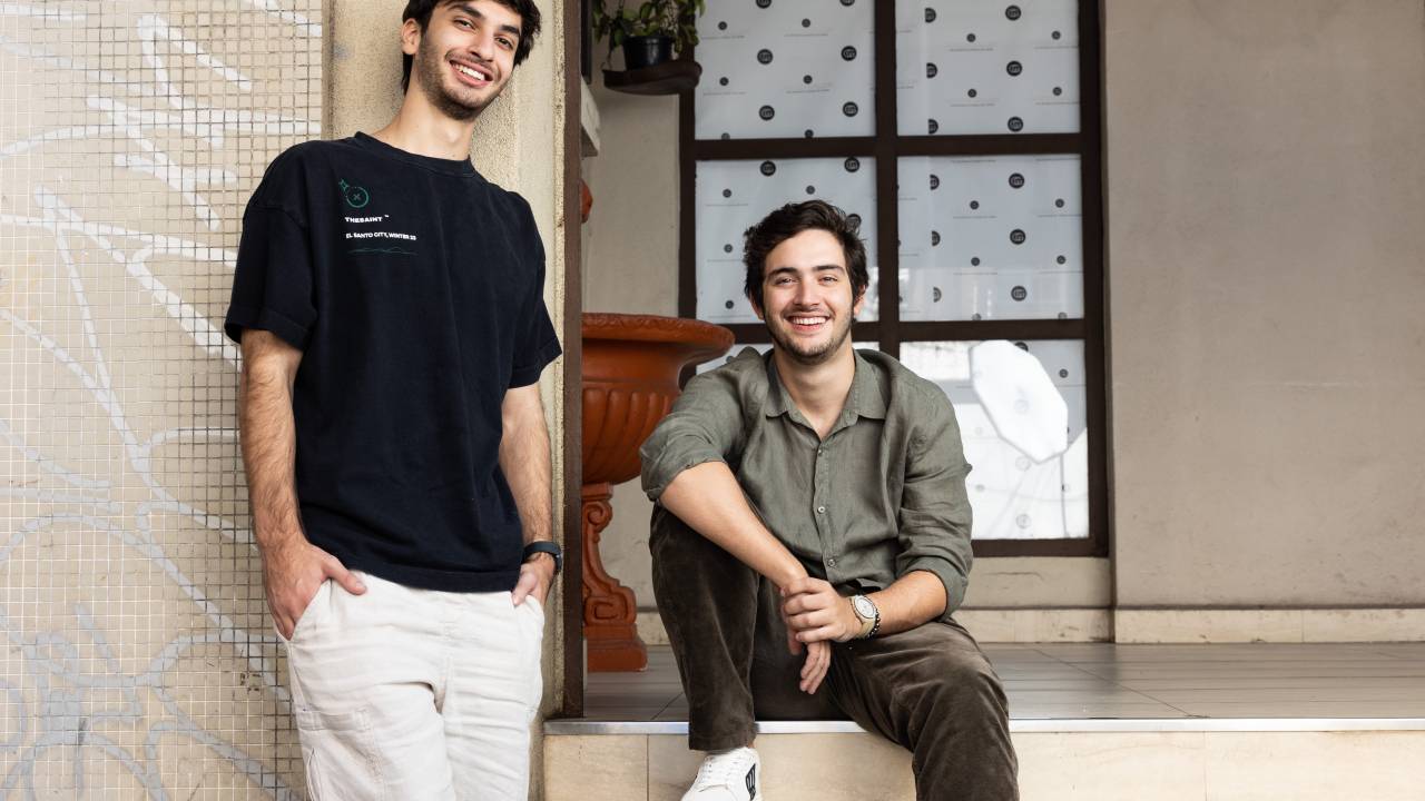 Dois jovens brancos posam encostados na fachada de galeria, um sentado e outro de pé