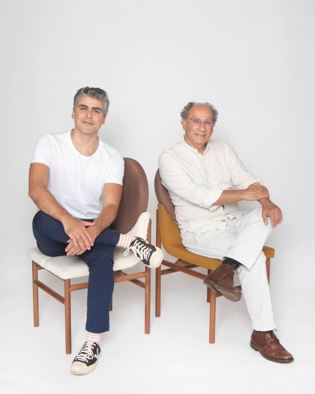 Dois homens grisalhos posam sentados em cadeiras e vestindo branco