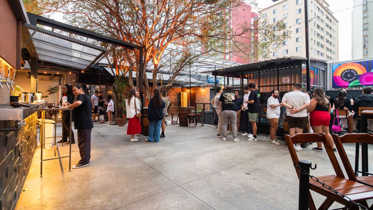 Ambiente da Croma Beer, com chão de cimento, árvore ao centro, chopeiras à esqueda e pessoas em pé à direito e ao fundo.