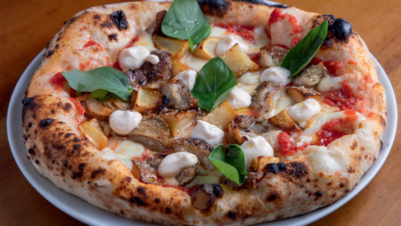 Pizza sobre prato de louça branca em cima de mesa de madeira com folhas de manjericão por cima.