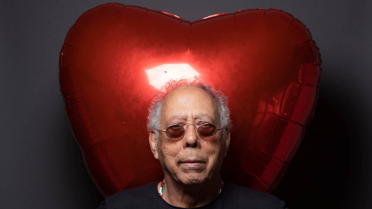 Imagem mostra homem grisalho com óculos vermelho, com balão em forma de coração ao fundo