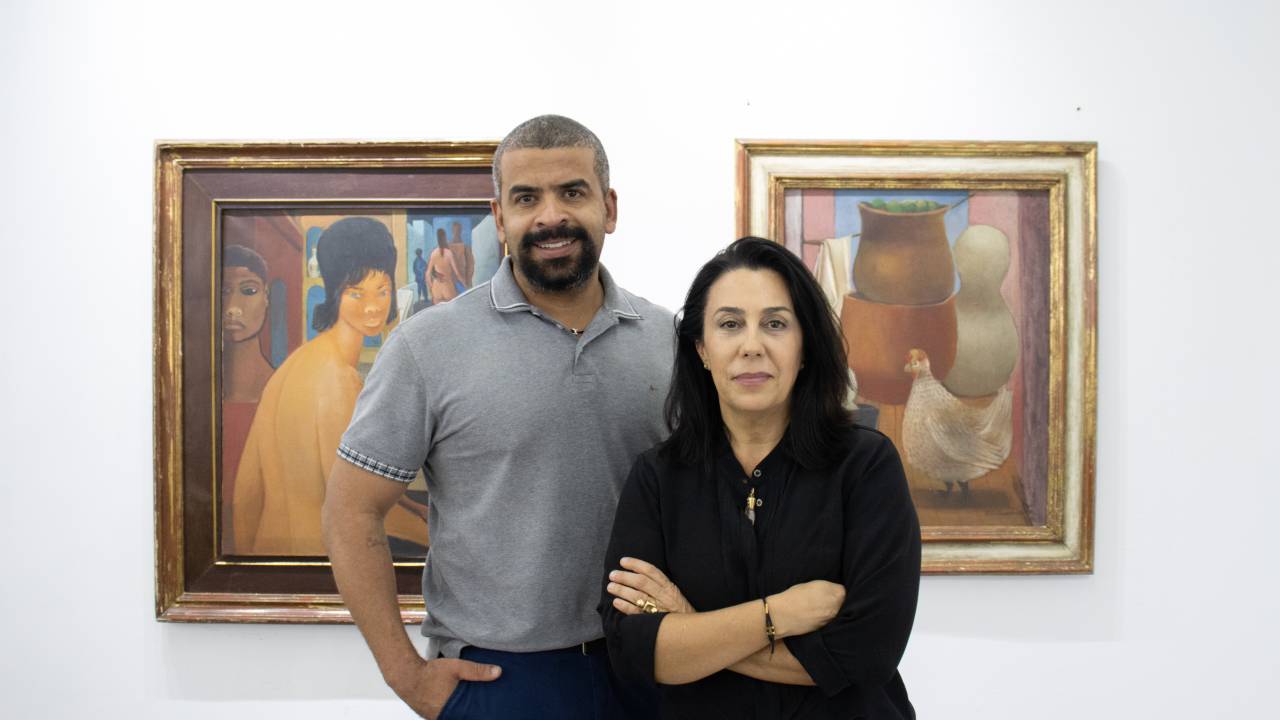 Homem negro e mulher branca posam em frente a dois quadros