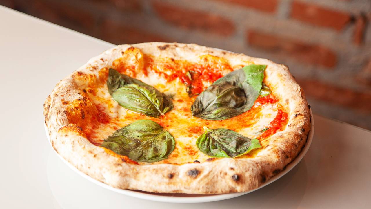 Pizza margherita individual em prato de louça com cobertura de queijo, molho de tomate e manjericão sobre uma mesa com paredes de tijolos à vista ao fundo
