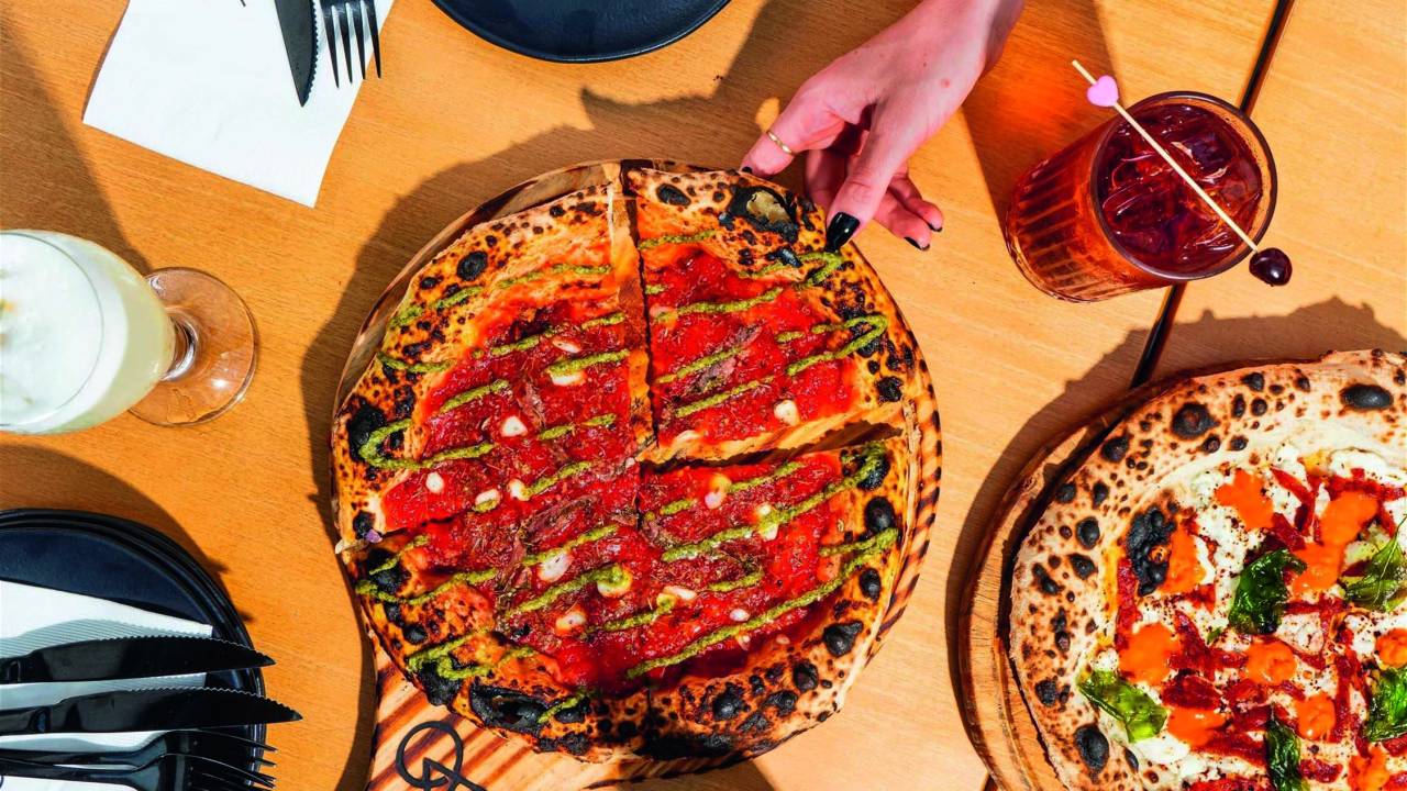 Foto aérea de mesa de madeira com pizza servida sobre suporte de madeira com drinks e mão puxando fatia.
