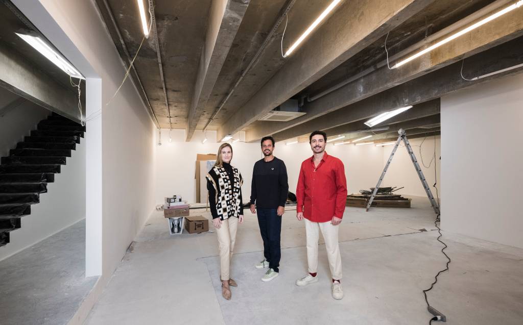 Três pessoas posam de pé em galpão em construção, a da direita vestindo camisa vermelha com calça branca e as demais com peças escuras