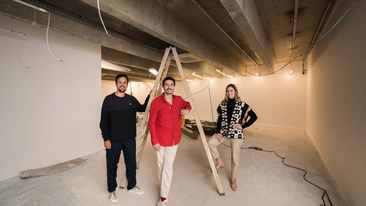 Três pessoas posam de pé em galpão em construção, a do meio vestindo camisa vermelha com calça branca e as demais com peças escuras